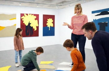 Foto von 3 Kindern und zwei Erwachsenen in einer Ausstellung: Auf dem Boden bunte Pappestücke, an den Wänden Bilder aus buntem Karton