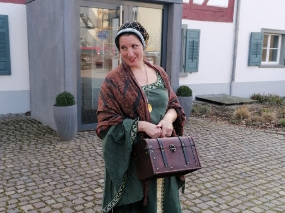 Foto von einer Frau in mittelalterlicher Kleidung mit kleinem Koffer in der Hand vor Fachwekhaus in Singen