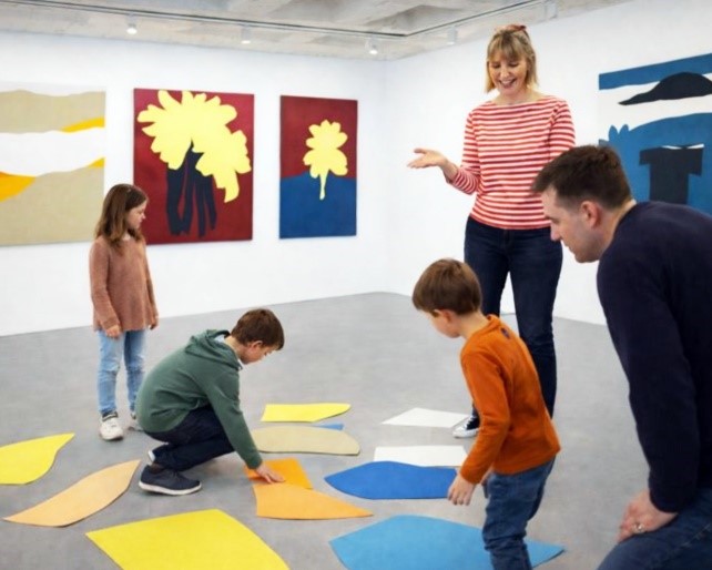 Foto von 3 Kindern und zwei Erwachsenen in einer Ausstellung: Auf dem Boden bunte Pappestücke, an den Wänden Bilder aus buntem Karton